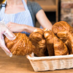 female baker s hand wearing plastic glove taking baked croissant from the basket