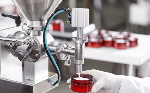 Hard mechanism of packing ready beauty cream into red container. A factory worker dressed in white lab gown and gloves holding red container under tap pouring ready cream. Production process of cream