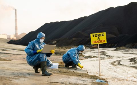 young-female-researcher-using-laptop-to-enter-data-about-description-of-toxic-water-in-front-of-board-announcing-danger-and-polluted-river